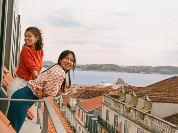 Two young women leaning on a balcony railing overlooking a coastal city with terracotta rooftops and a body of water in the background under a partly cloudy sky.
