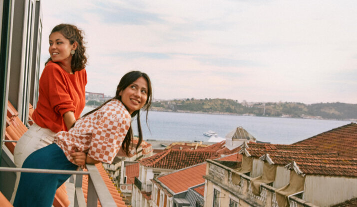 Two young women leaning on a balcony railing overlooking a coastal city with terracotta rooftops and a body of water in the background under a partly cloudy sky.