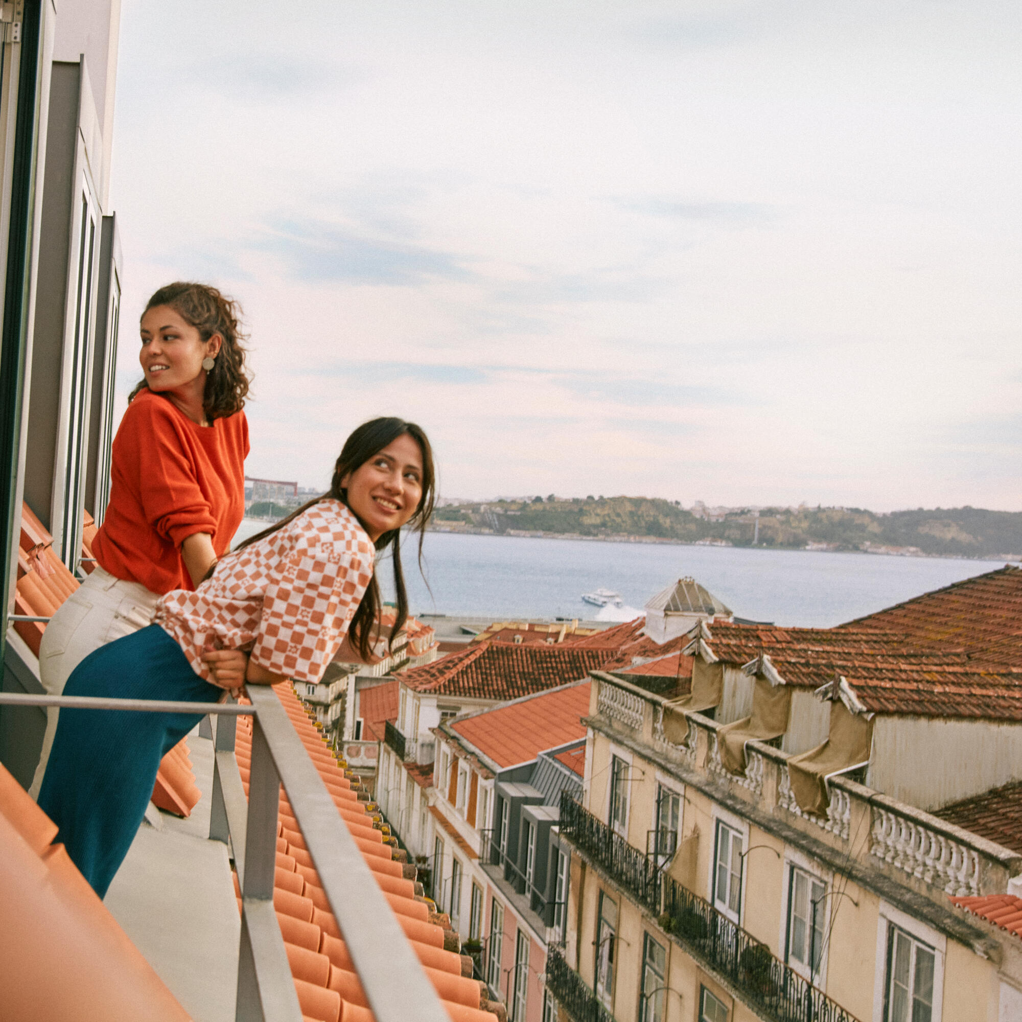 Two young women leaning on a balcony railing overlooking a coastal city with terracotta rooftops and a body of water in the background under a partly cloudy sky.