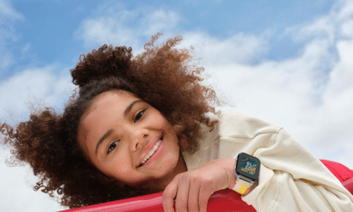 Smiling young person with curly hair leaning on red playground bars against a partly cloudy blue sky, wearing a white sweater with smiley face patches and a smartwatch with a colorful band.
