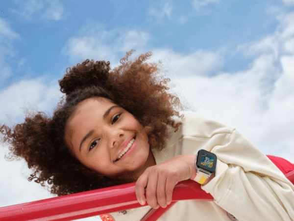 Smiling young person with curly hair leaning on red playground bars against a partly cloudy blue sky, wearing a white sweater with smiley face patches and a smartwatch with a colorful band.