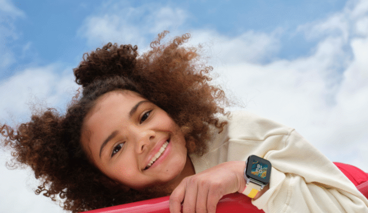 Smiling young person with curly hair leaning on red playground bars against a partly cloudy blue sky, wearing a white sweater with smiley face patches and a smartwatch with a colorful band.