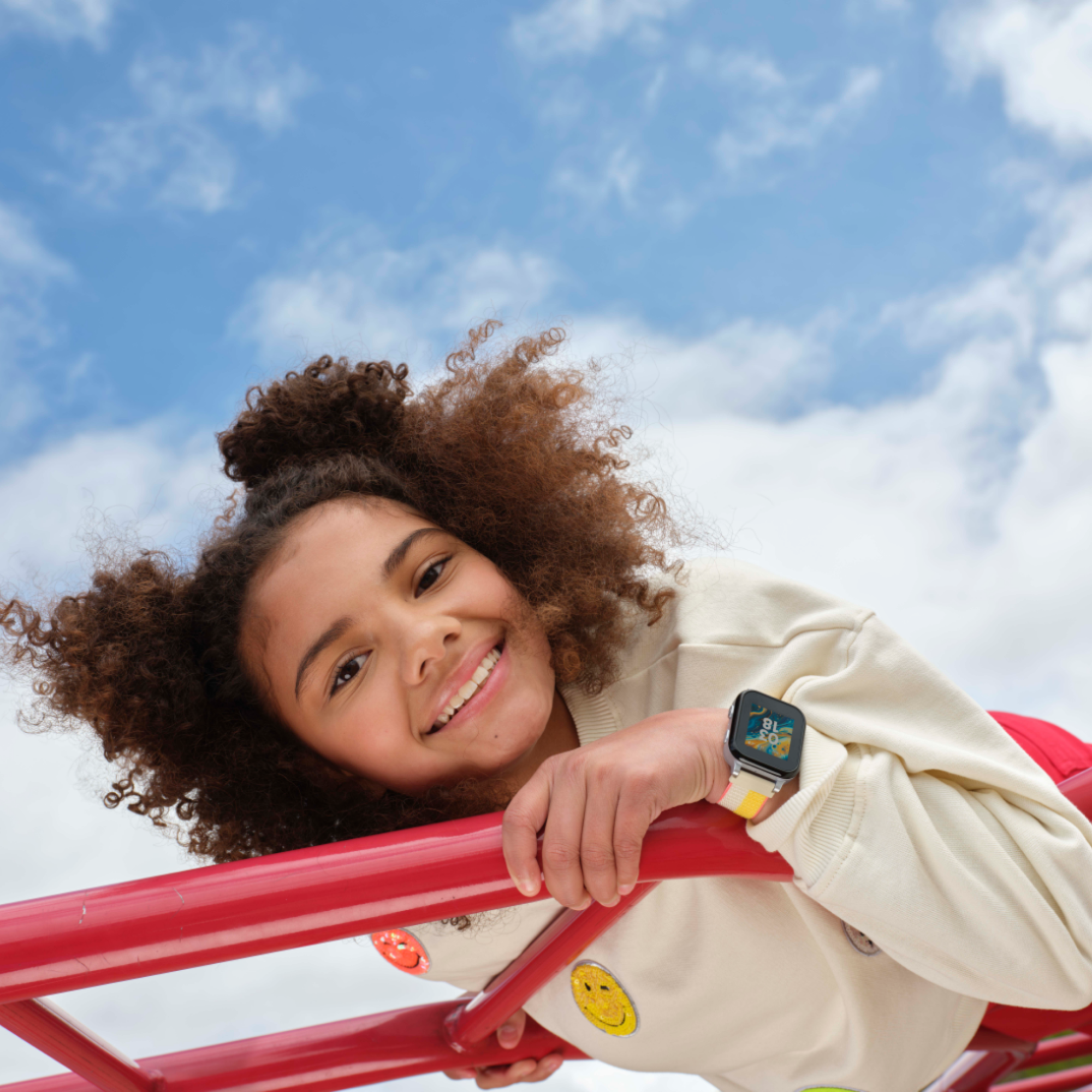 Smiling young person with curly hair leaning on red playground bars against a partly cloudy blue sky, wearing a white sweater with smiley face patches and a smartwatch with a colorful band.