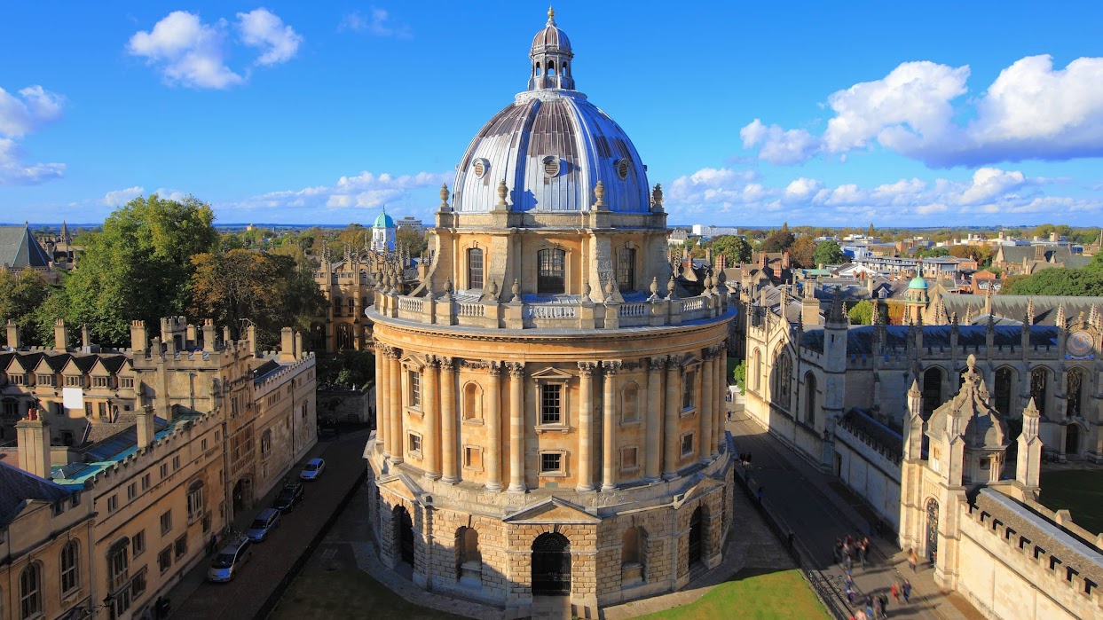 The Radcliffe Camera, a large circular stone building with a prominent dome, set among historic university buildings under a bright blue sky with scattered clouds.