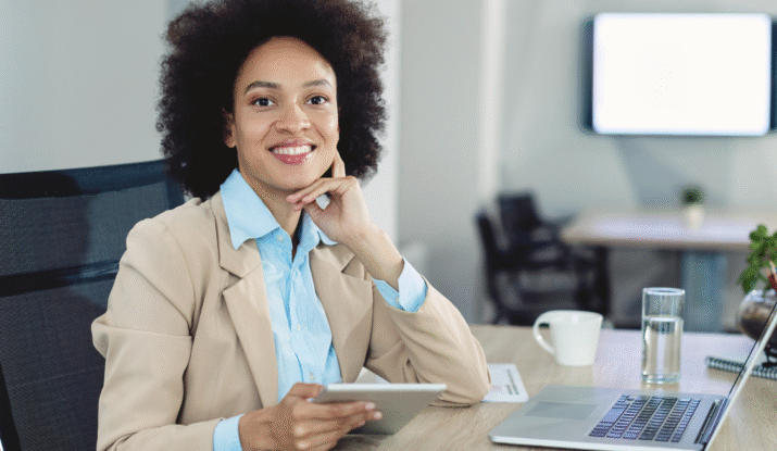 Smiling woman with curly hair wearing a beige blazer and blue shirt sitting at a desk in an office, holding a tablet with an open laptop, a glass of water, and a white coffee mug in front of her