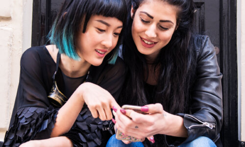 Two young women sitting closely together in front of a black door, smiling and looking at a smartphone one holds, with one woman having black hair with teal tips and the other with long dark hair styled in two buns, wearing casual outfits including a black leather jacket and layered black top.
