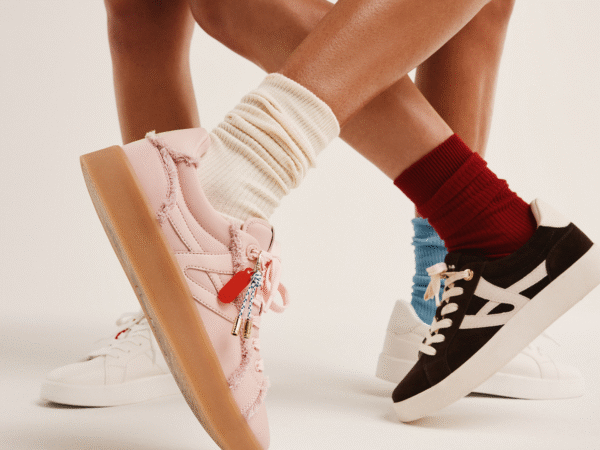 Close-up of two pairs of legs wearing colorful sneakers and socks, one in pink sneakers with cream socks and the other in black sneakers with red socks, against a plain light background.