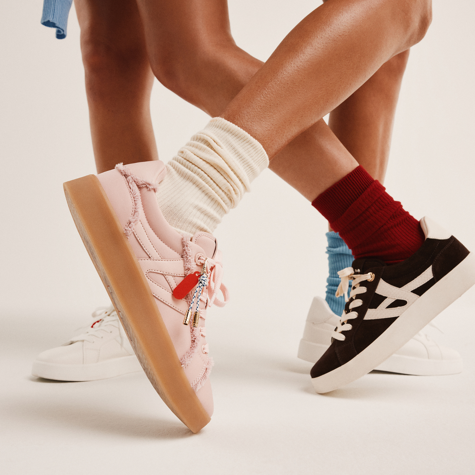 Close-up of two pairs of legs wearing colorful sneakers and socks, one in pink sneakers with cream socks and the other in black sneakers with red socks, against a plain light background.