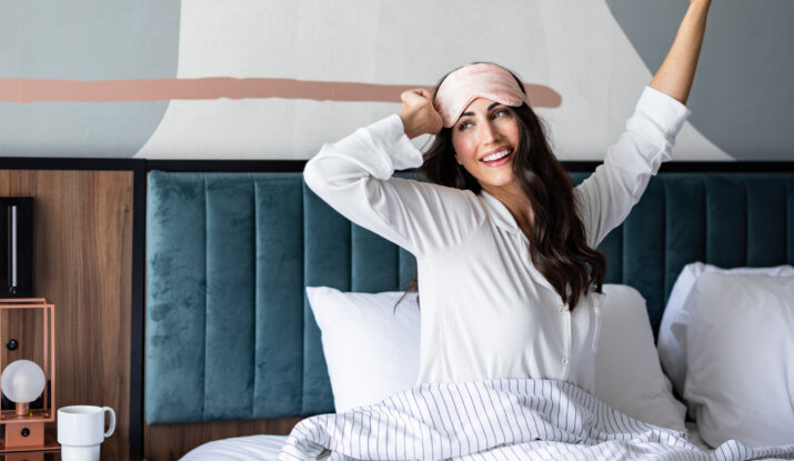 Woman in white pajamas wearing a pink sleep mask on her forehead, sitting up in bed stretching with a smile, covered by a white blanket with thin gray stripes, with a stylish dark teal headboard and wooden nightstand beside her holding a white mug and a modern lamp.