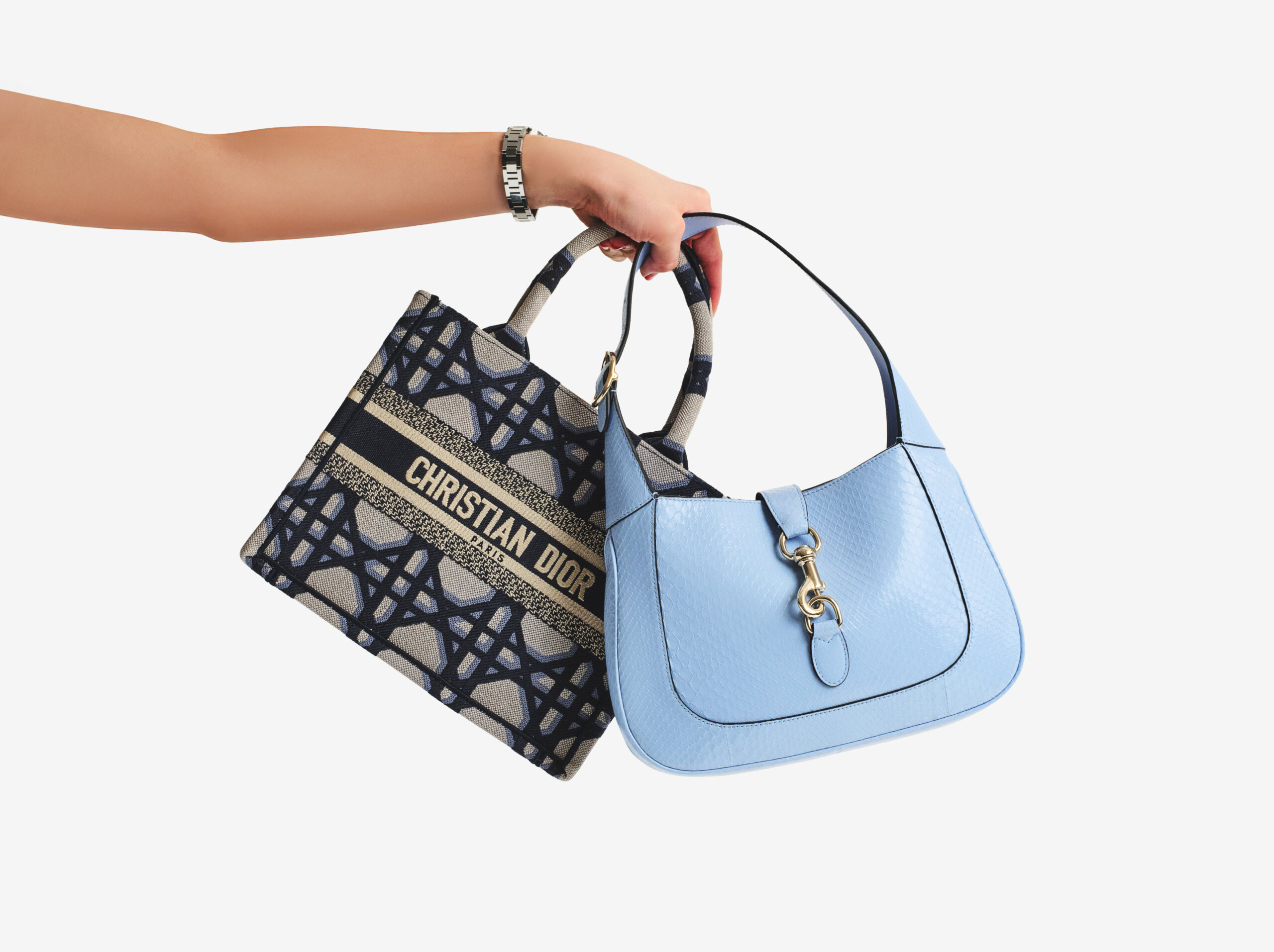A hand wearing a silver watch holds two handbags: a blue leather handbag with a textured pattern and a gold clasp, and a beige and navy Christian Dior tote bag with a geometric pattern and the brand name on it.
