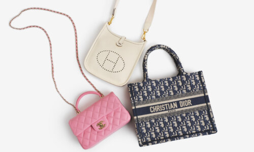 Three designer handbags on a white background: a white Hermes crossbody bag with perforated logo, a pink Chanel quilted leather handbag with chain strap, and a blue Christian Dior logo tote bag.