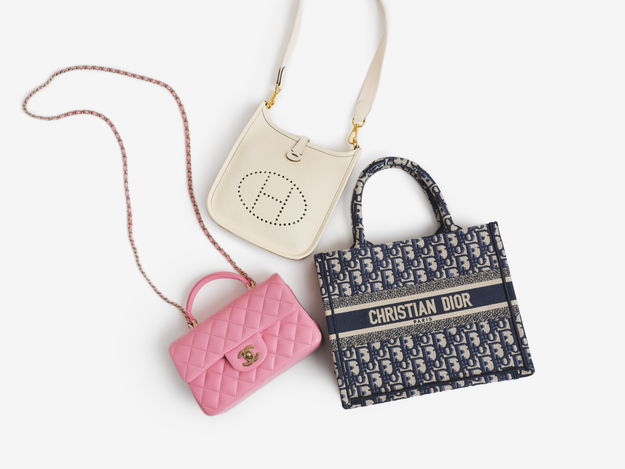 Three designer handbags on a white background: a white Hermes crossbody bag with perforated logo, a pink Chanel quilted leather handbag with chain strap, and a blue Christian Dior logo tote bag.
