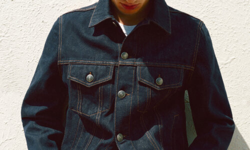 Young man wearing a dark denim jacket with prominent stitching, looking down against a textured white wall background.