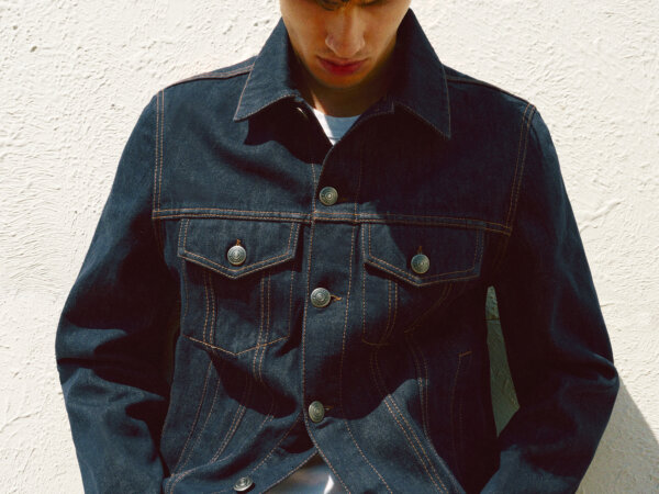 Young man wearing a dark denim jacket with prominent stitching, looking down against a textured white wall background.