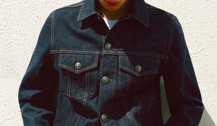 Young man wearing a dark denim jacket with prominent stitching, looking down against a textured white wall background.