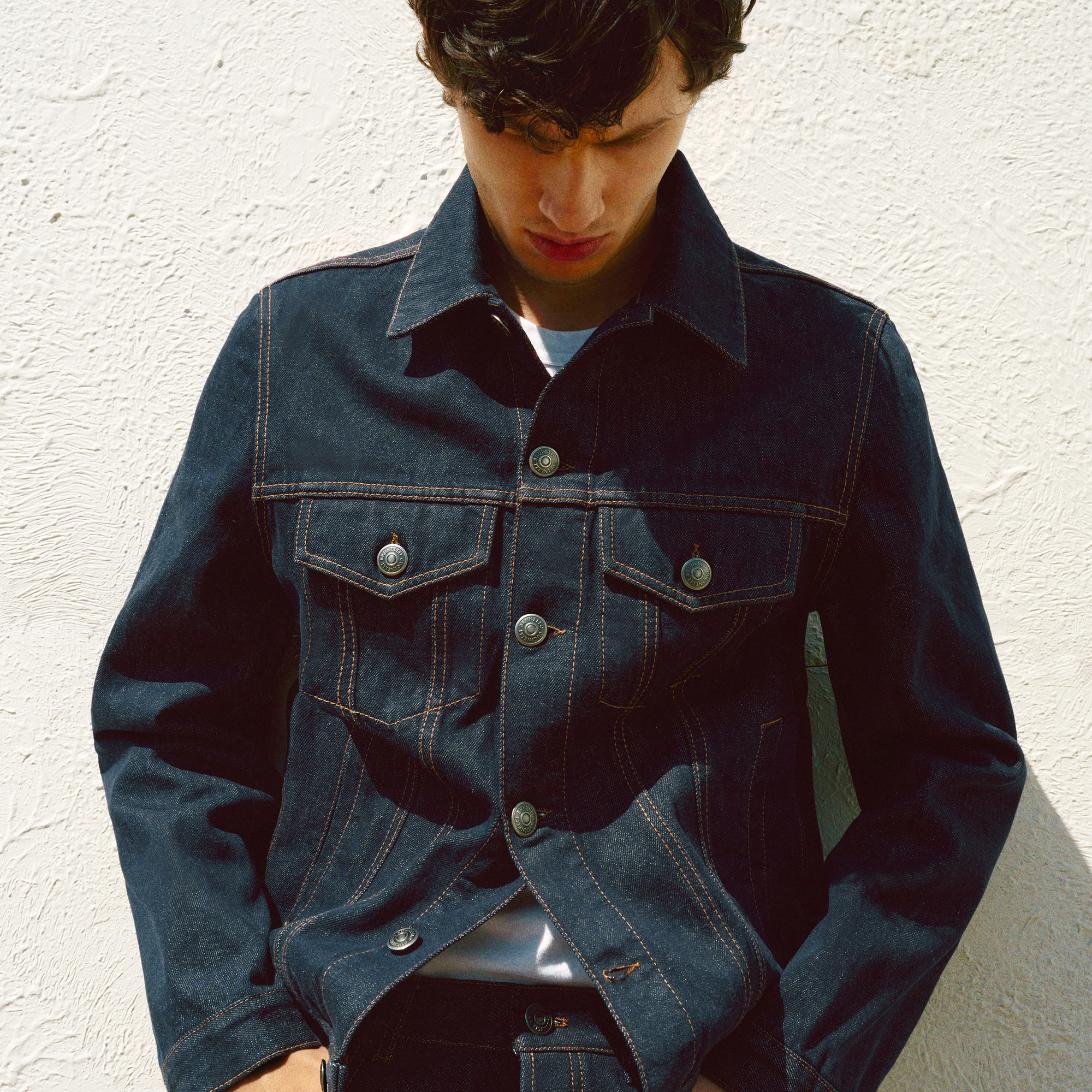 Young man wearing a dark denim jacket with prominent stitching, looking down against a textured white wall background.