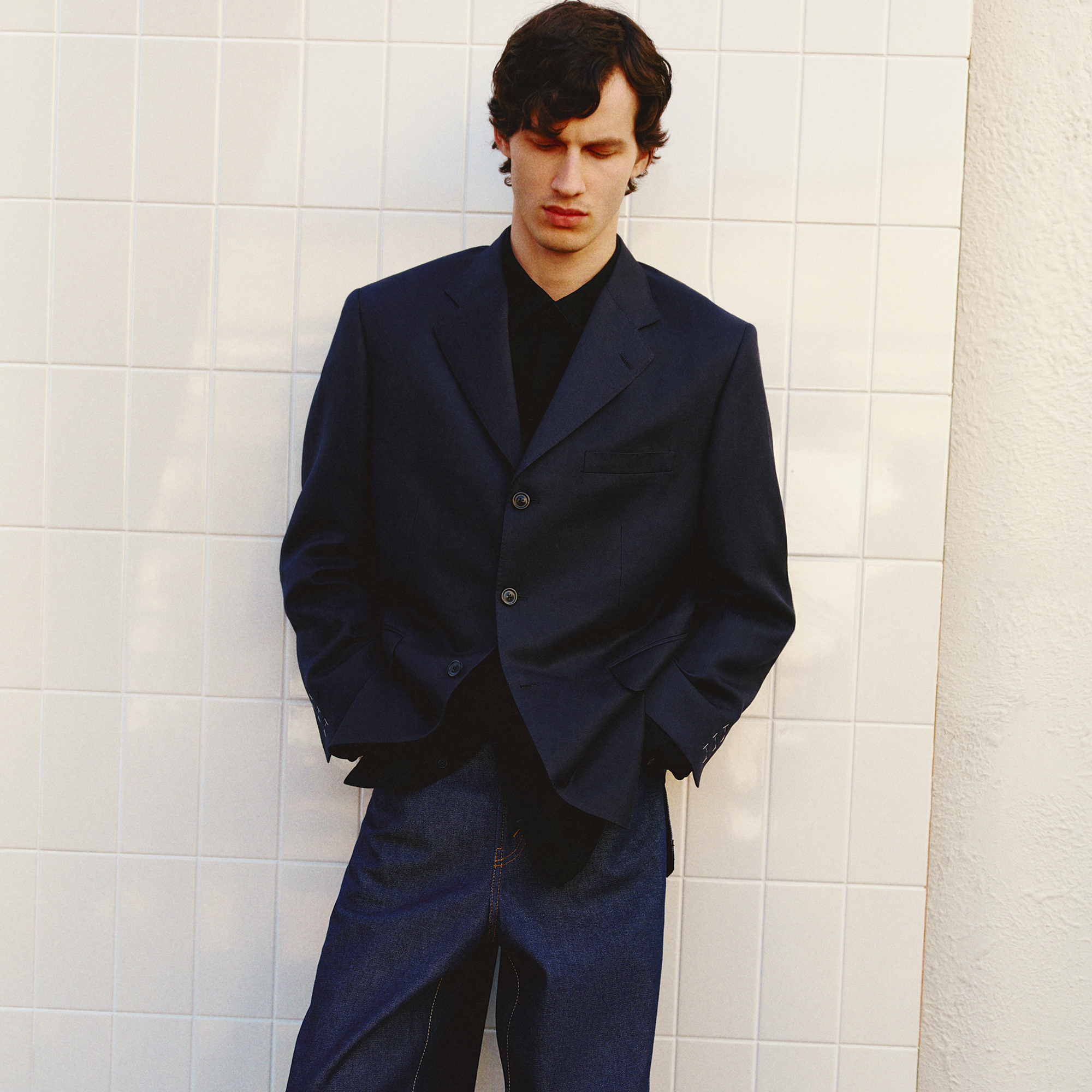 Young man with curly brown hair wearing a navy blazer, black shirt, and dark blue jeans, standing against a white tiled wall with his hands in his pockets and eyes closed.