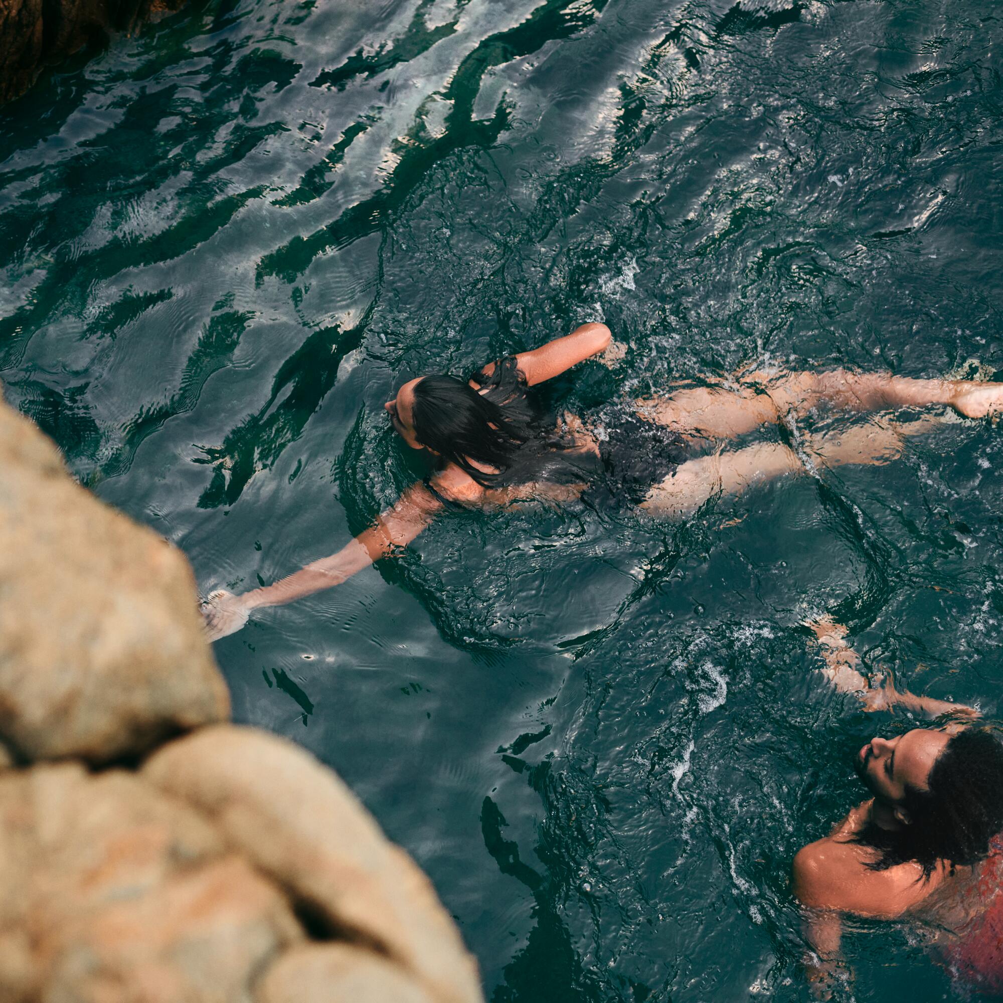 Two people swimming in dark green water near large rocks, one reaching out to touch the rocks while the other swims nearby.