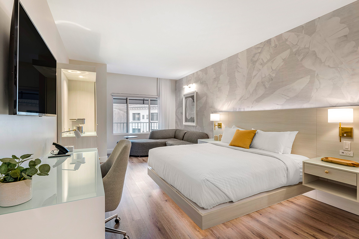 Modern hotel room with a large bed featuring white linens and a yellow accent pillow, a grey L-shaped sofa near a window, wooden flooring, a desk with a chair and phone, a large flat-screen TV mounted on the wall, and wall lamps providing soft lighting against a light-colored patterned wallpaper.