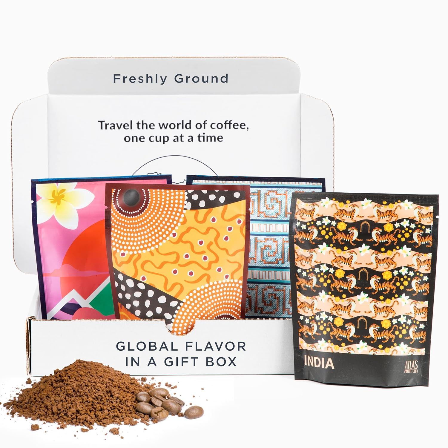 A white gift box labeled Global Flavor in a Gift Box containing four colorful coffee bags with artistic designs, with coffee grounds and beans in front of the box and text inside the box reading 'Freshly Ground' and 'Travel the world of coffee, one cup at a time'.