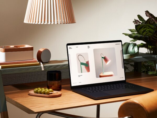 Wooden desk with a laptop displaying images of a modern desk lamp, next to a tray with green grapes, a brown glass, a stack of books, a wooden organizer, a round speaker, green headphones, and a large houseplant in the background under a hanging lamp shade.
