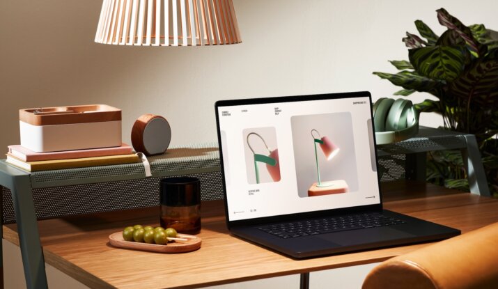 Wooden desk with a laptop displaying images of a modern desk lamp, next to a tray with green grapes, a brown glass, a stack of books, a wooden organizer, a round speaker, green headphones, and a large houseplant in the background under a hanging lamp shade.