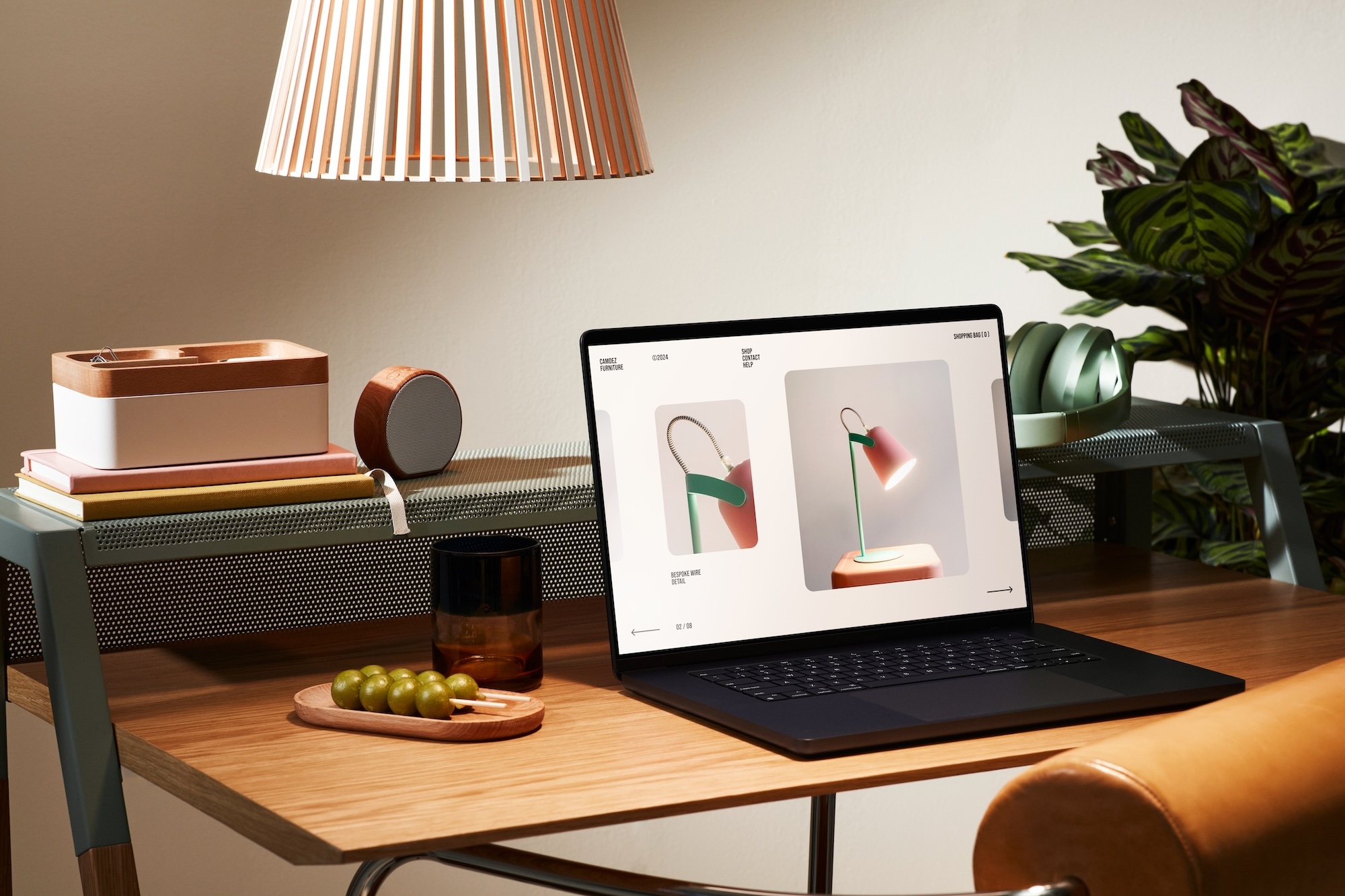 Wooden desk with a laptop displaying images of a modern desk lamp, next to a tray with green grapes, a brown glass, a stack of books, a wooden organizer, a round speaker, green headphones, and a large houseplant in the background under a hanging lamp shade.