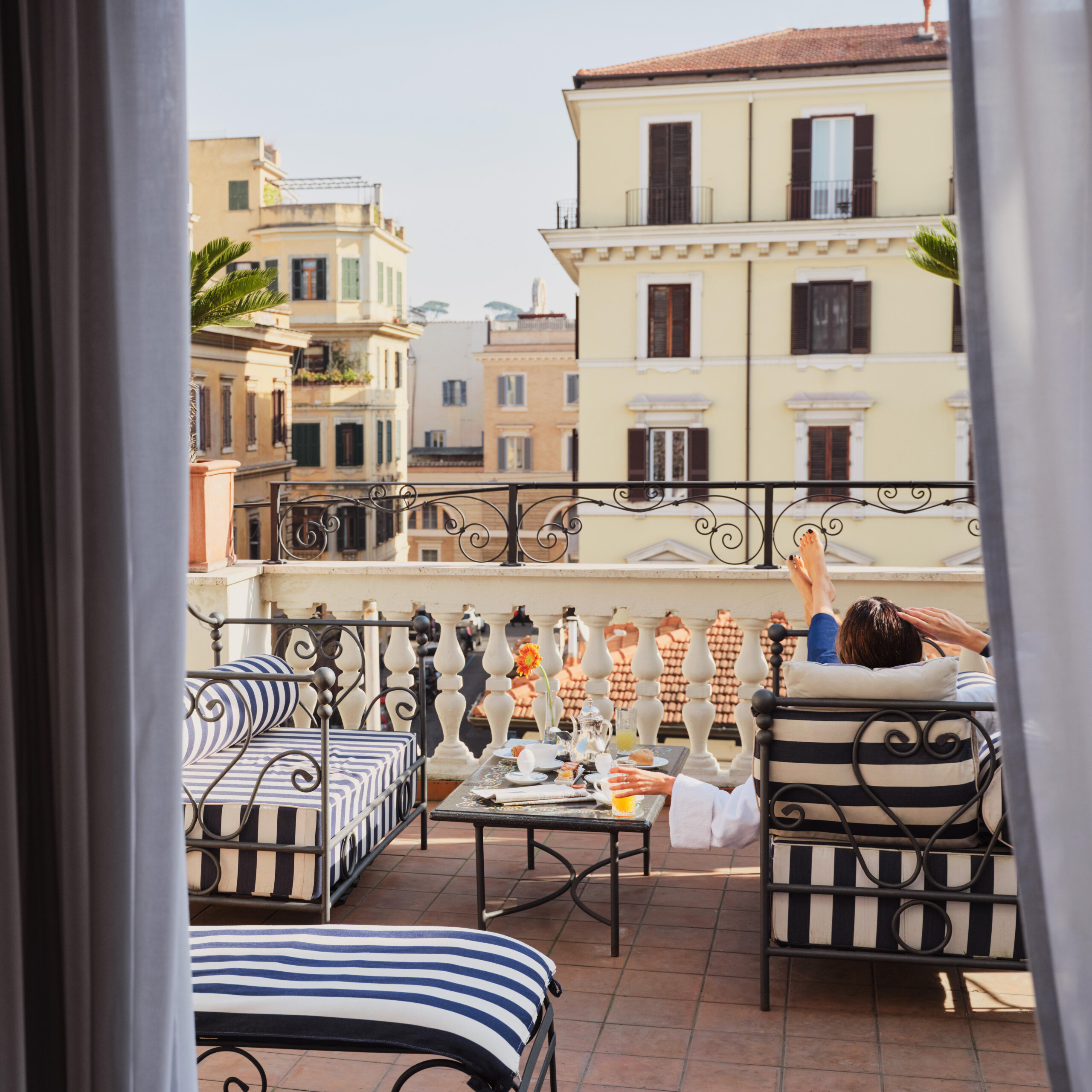 Balcony with black wrought iron furniture featuring blue and white striped cushions, a table with breakfast items, and a person reclining on a chair with feet up, overlooking classical buildings in sunlight.
