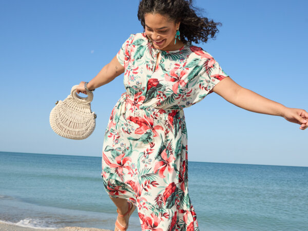 A woman in a floral dress joyfully balances on one foot while holding a woven handbag on a sandy beach with the ocean and clear blue sky in the background.