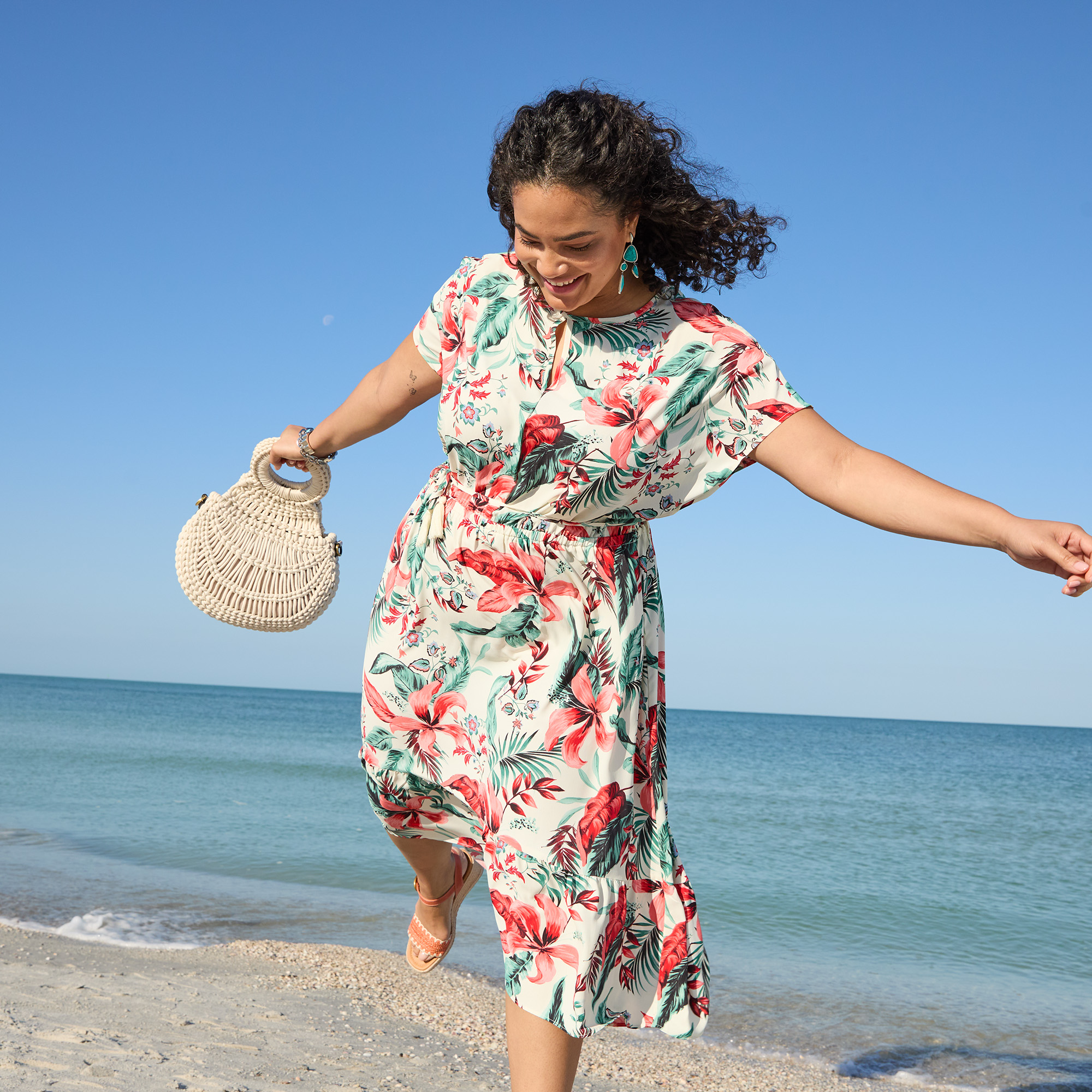 A woman in a floral dress joyfully balances on one foot while holding a woven handbag on a sandy beach with the ocean and clear blue sky in the background.
