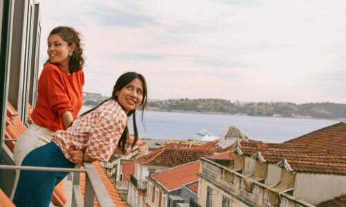Two women leaning on a balcony railing overlooking tiled rooftops, water, and distant hills under a partly cloudy sky, one wearing a red top and the other a checkered shirt.