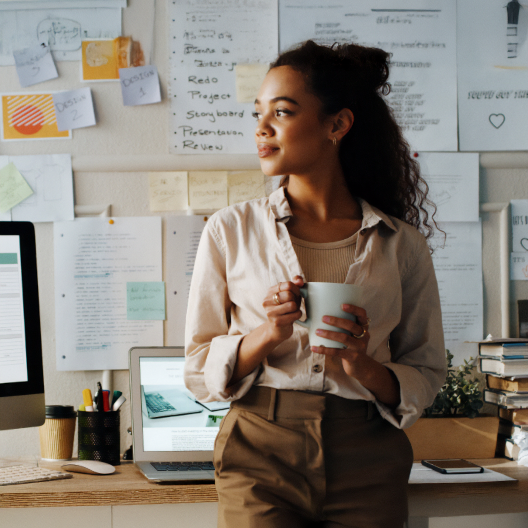 A woman with curly hair holding a mug, standing thoughtfully in front of a work desk with a laptop, computer monitor, stationery, and papers pinned on the wall behind her.