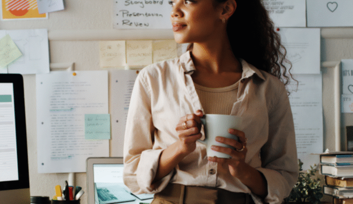 Young woman with curly hair holding a mug, standing and looking to the side in a workspace with a laptop, computer monitor, pens, books, and a wall covered with pinned papers and notes behind her.