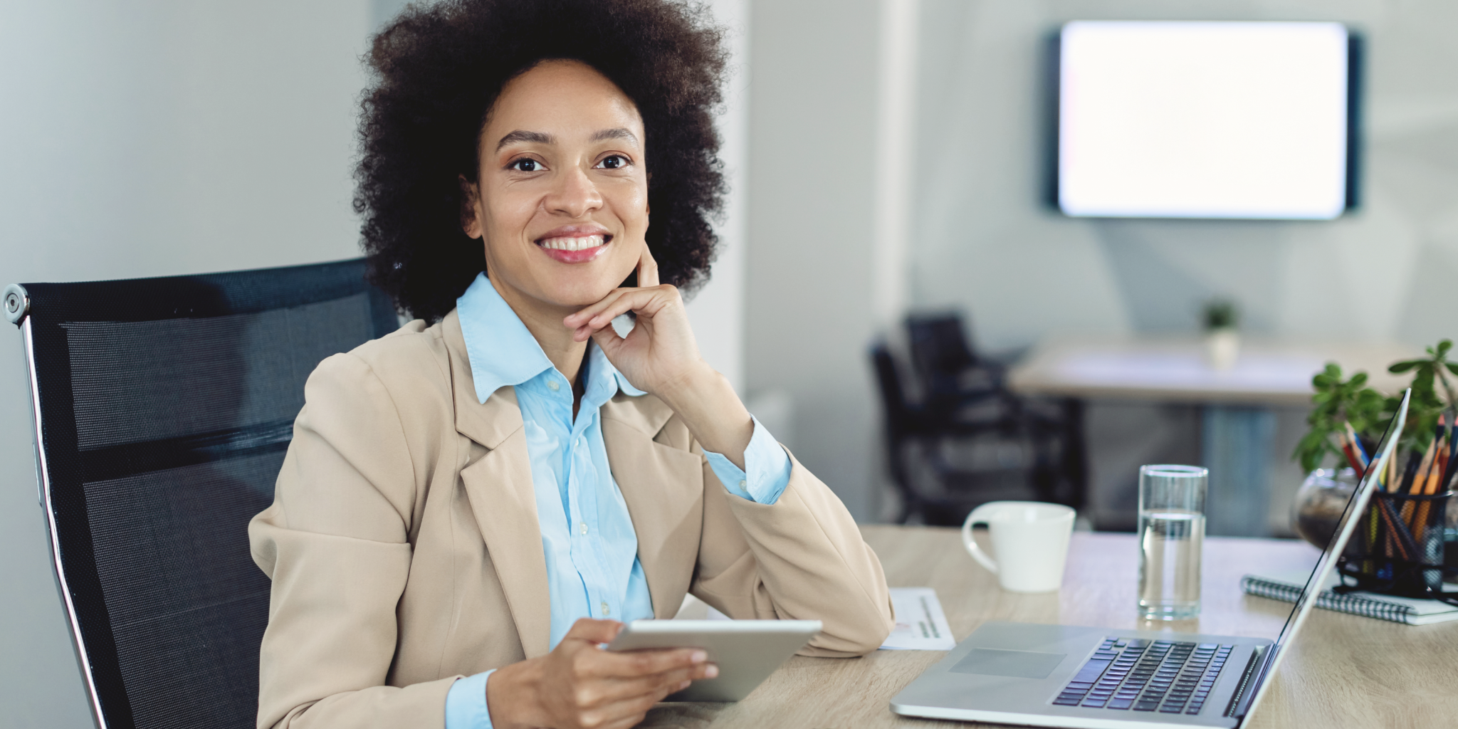 Smiling woman with curly hair sitting at a desk in an office, holding a tablet, with a laptop, glass of water, coffee cup, and pencil holder on the desk, and a blurred screen in the background.