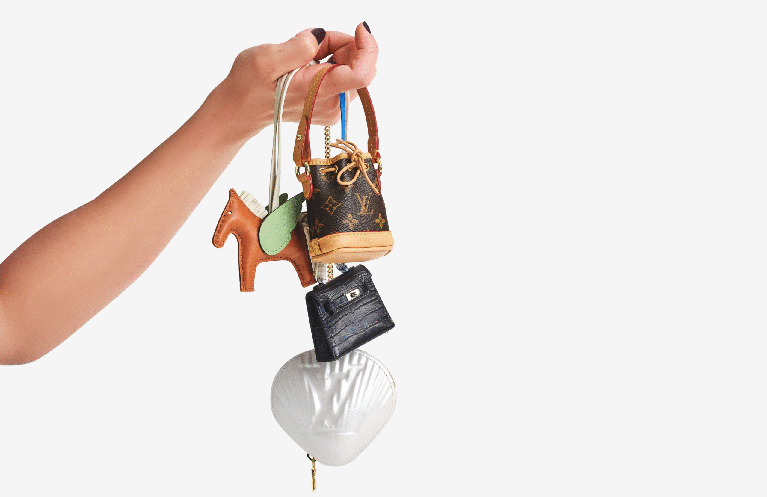Hand holding four luxury keychains: a brown leather horse with green wings, a small brown Louis Vuitton handbag, a small black crocodile leather handbag, and a white seashell-shaped item with the Louis Vuitton logo embossed on it.