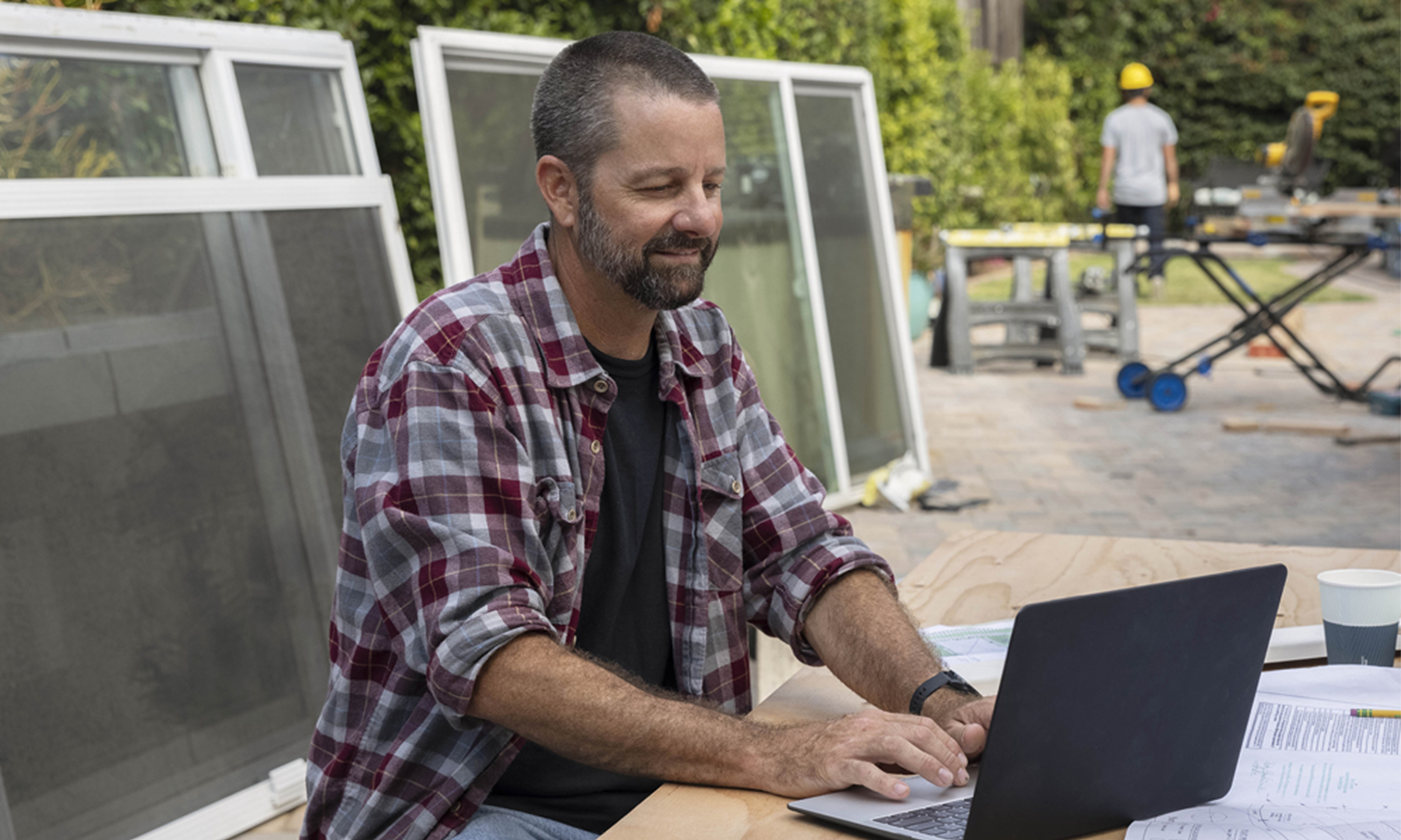 Bearded man wearing a plaid shirt working outdoors at a laptop with window frames and construction tools nearby