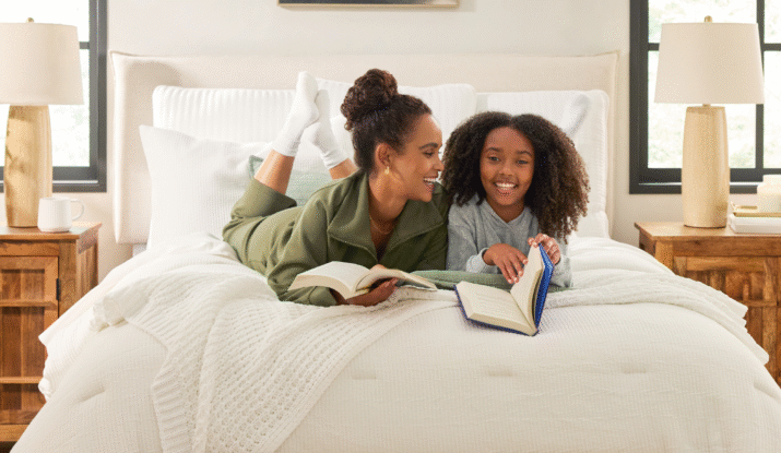 Two people lying on a bed reading books, one with hair in a bun wearing a green outfit and the other with curly hair wearing a gray sweater, in a bright bedroom with wooden nightstands, lamps, and windows on either side of the bed.
