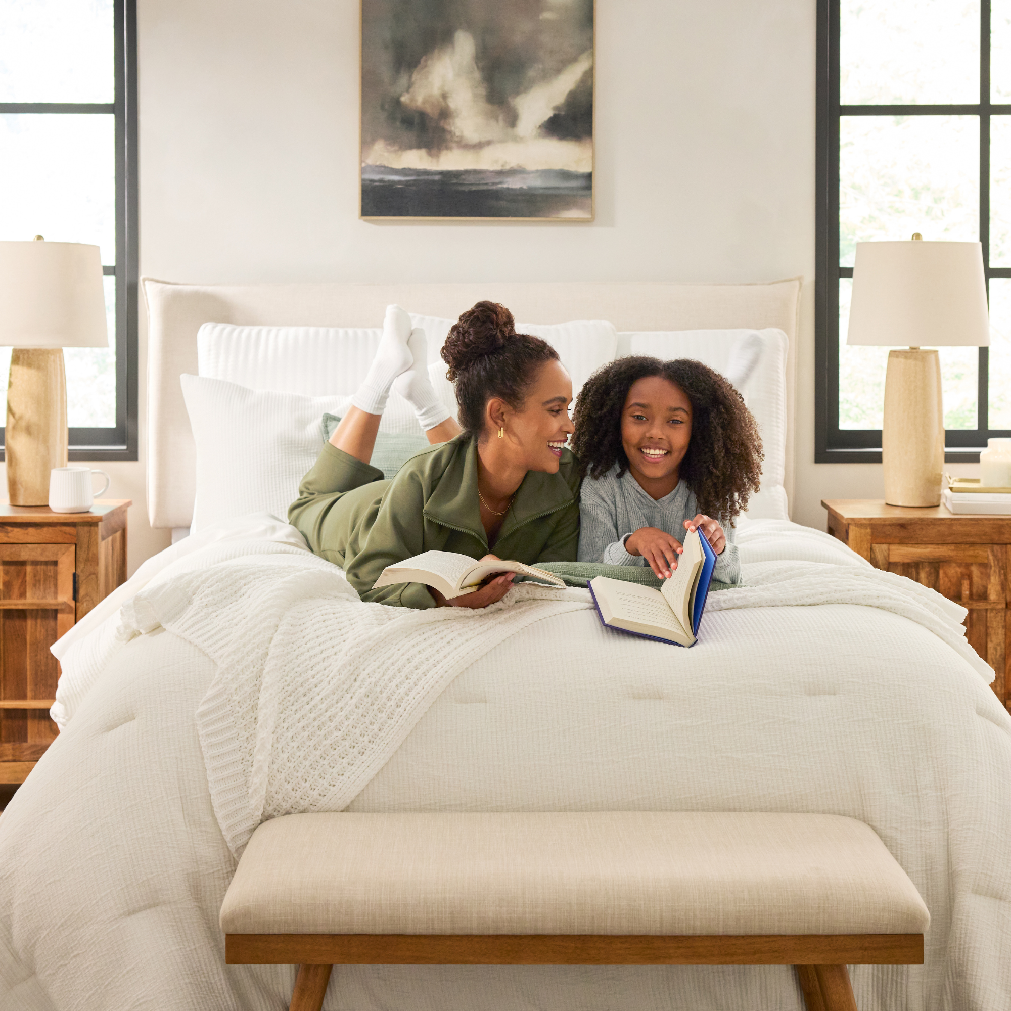 Two people lying on a bed reading books, one with hair in a bun wearing a green outfit and the other with curly hair wearing a gray sweater, in a bright bedroom with wooden nightstands, lamps, and windows on either side of the bed.