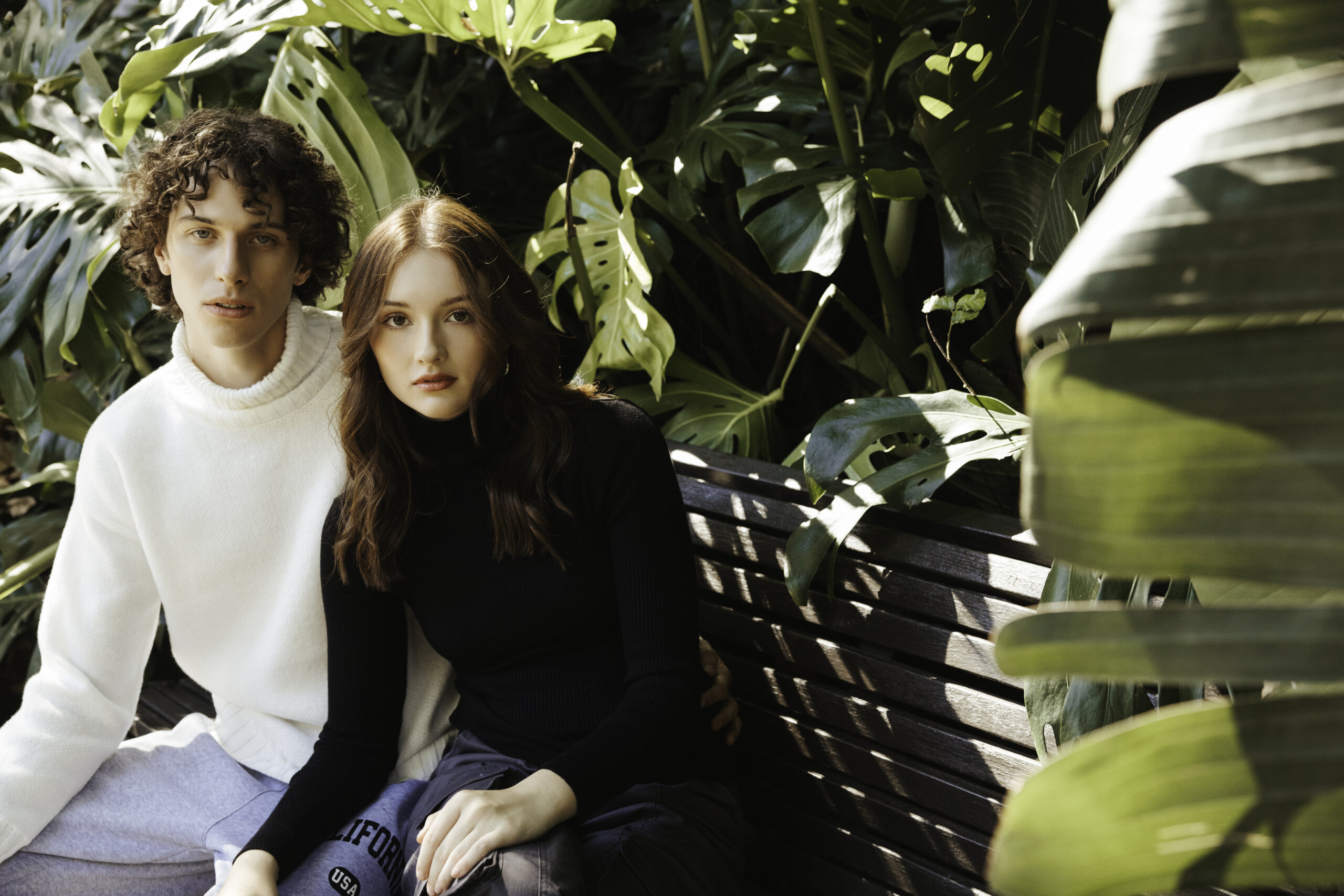 Young man with curly hair wearing a white sweater and a young woman with long brown hair wearing a black turtleneck sitting closely together on a wooden bench surrounded by large green tropical leaves.
