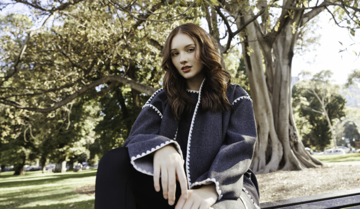 Young woman with long reddish-brown hair wearing a dark gray coat with white trim, sitting on a park bench with trees and greenery in the background on a sunny day.