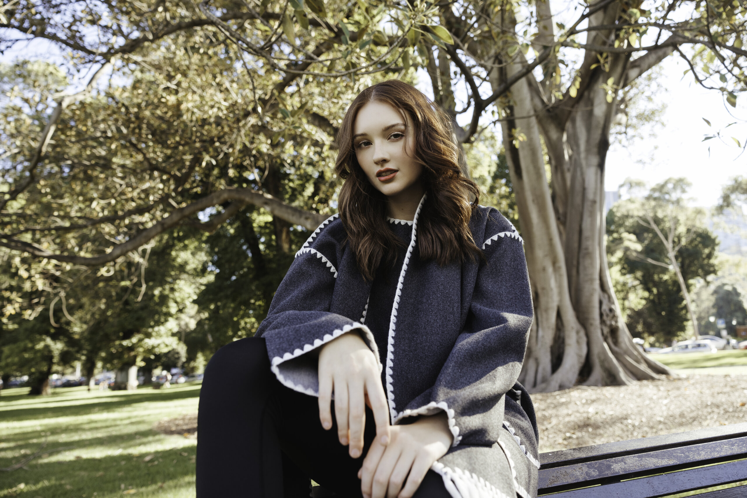 Young woman with long reddish-brown hair wearing a dark gray coat with white trim, sitting on a park bench with trees and greenery in the background on a sunny day.
