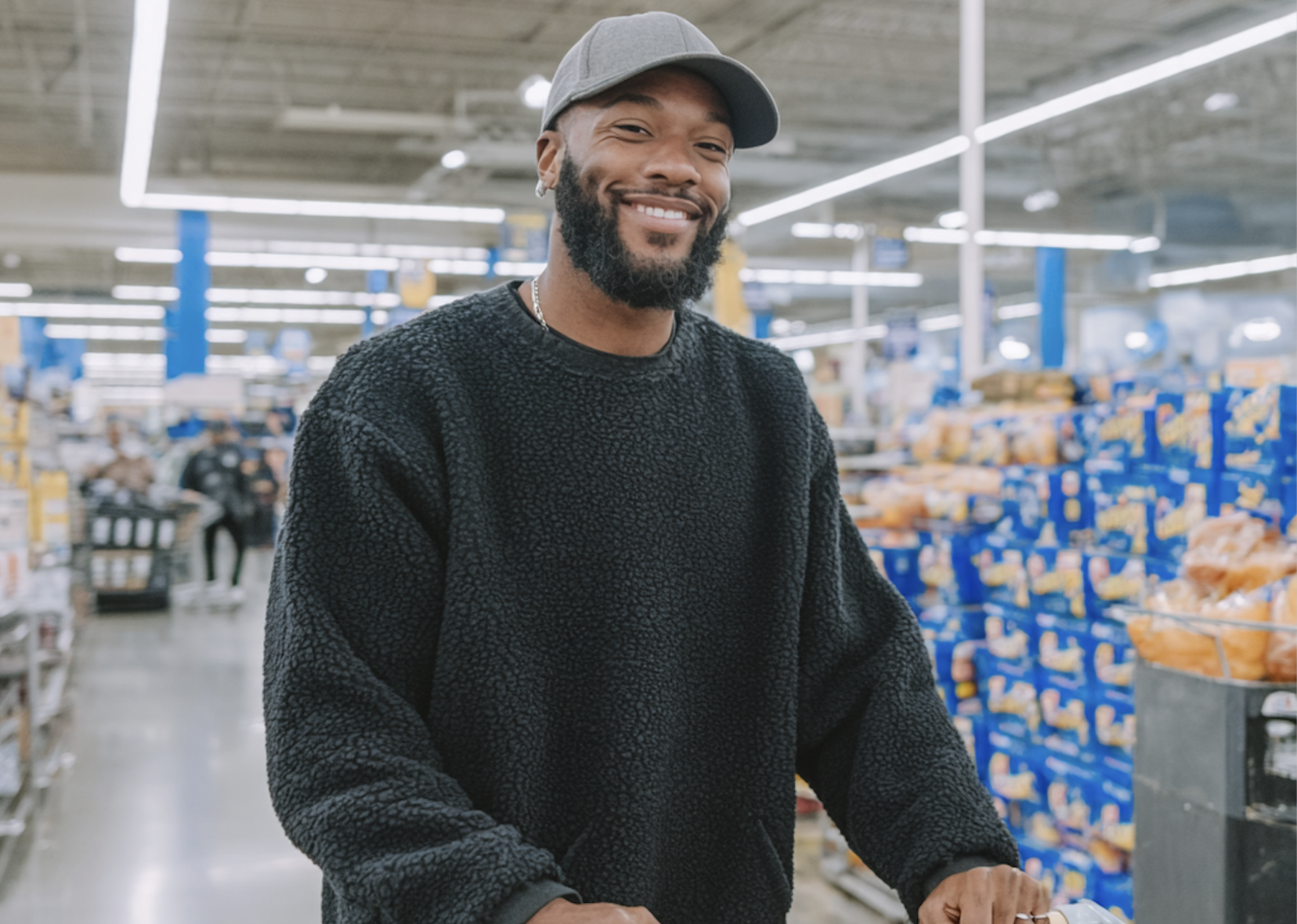 Smiling man wearing a gray baseball cap and a black textured sweater pushing a shopping cart in a brightly lit grocery store aisle.