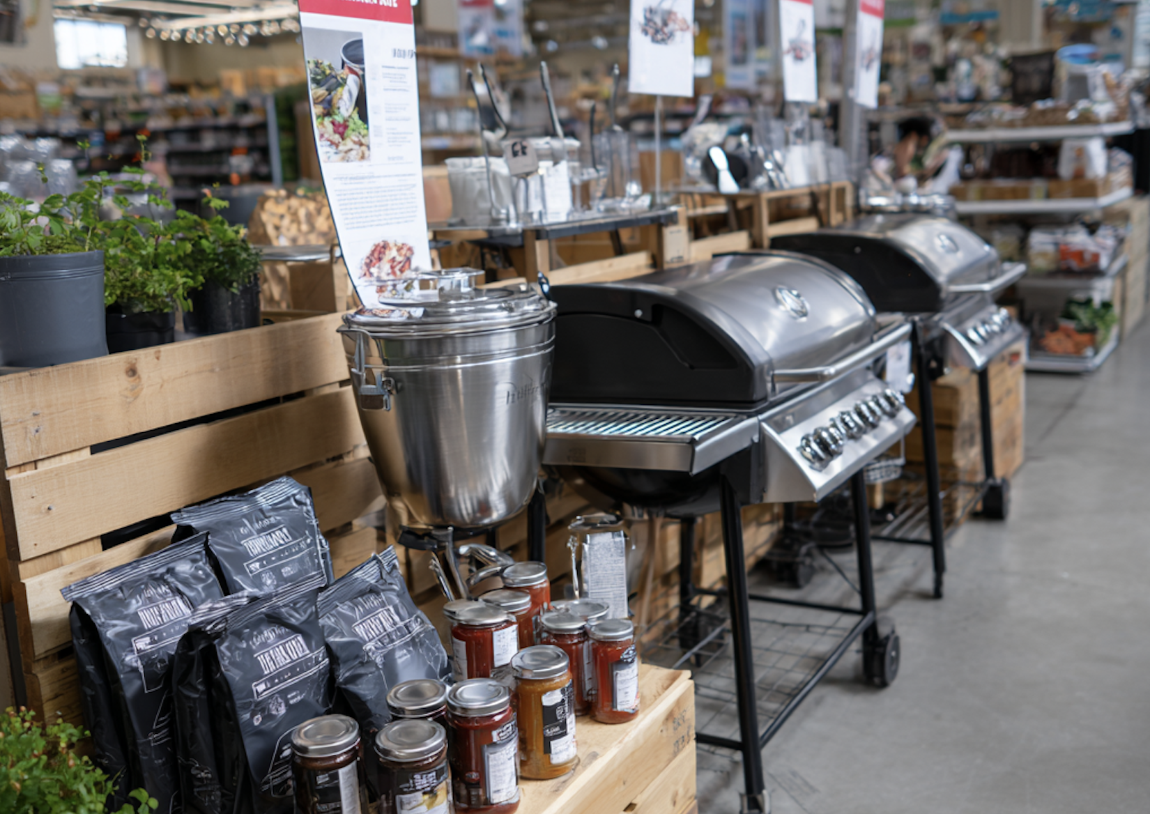 Store display with two stainless steel gas grills, a metal container, several jars of preserves, potted plants, and bags of charcoal arranged on and beside wooden crates and shelves.