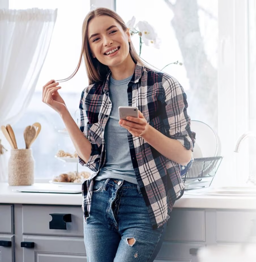 Young woman smiling and holding a smartphone while standing in a bright kitchen, wearing a plaid shirt over a blue top and ripped jeans.
