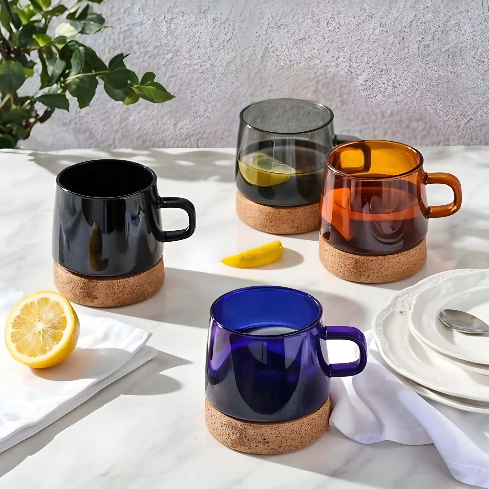 Four colored glass mugs with cork bottoms on a white surface, surrounded by a half lemon on a white napkin, a yellow chili pepper, and stacked white plates with a spoon, with green leaves and a textured gray wall in the background.