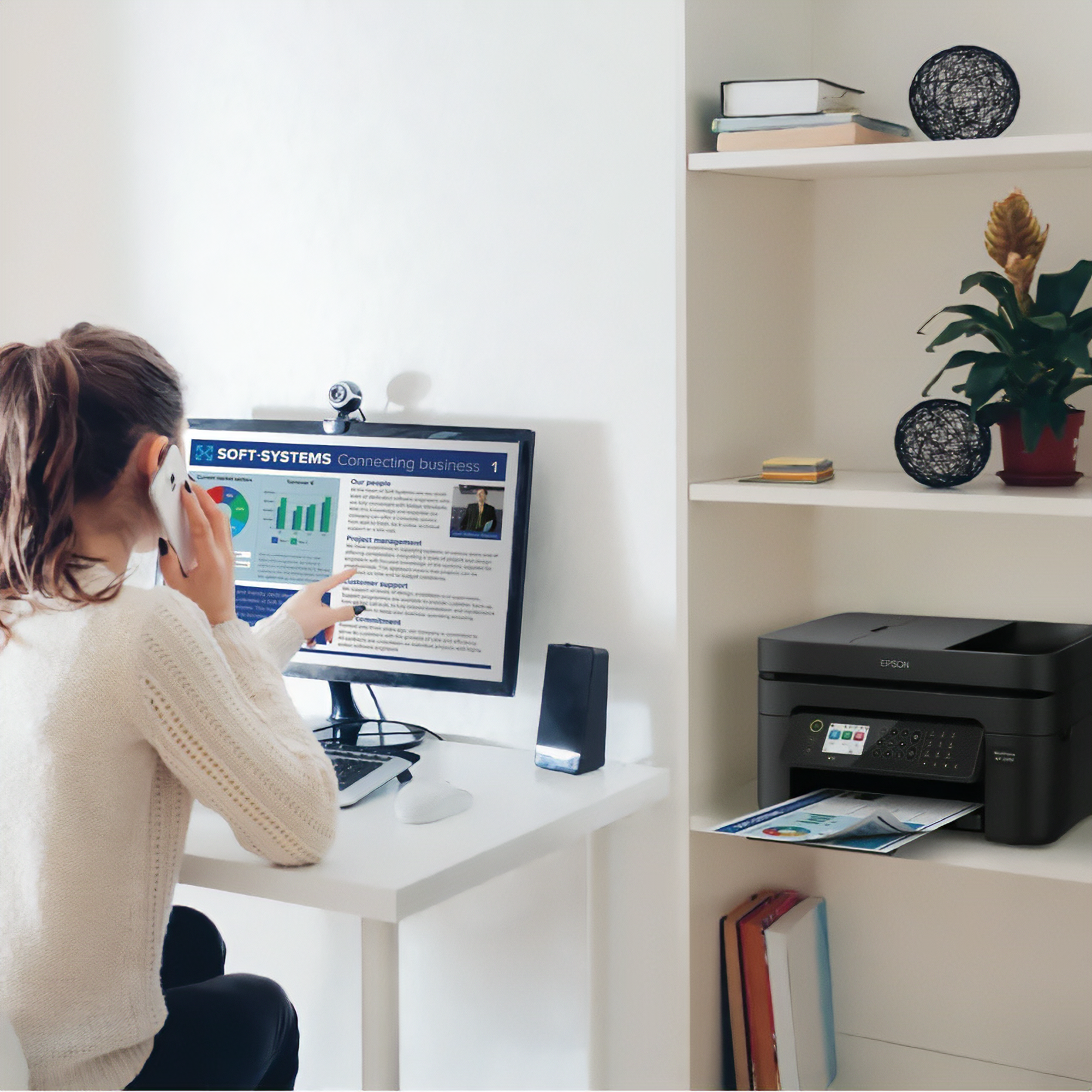 Woman in white sweater sitting at desk talking on phone and pointing at computer screen with business information, next to a shelf holding books, decorative items, a plant, and a black Epson printer printing documents.