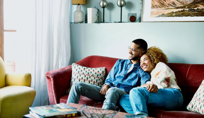 Smiling couple sitting closely together on a red couch in a cozy living room with patterned pillows, a yellow armchair, and a coffee table with books and a decorative wire basket.