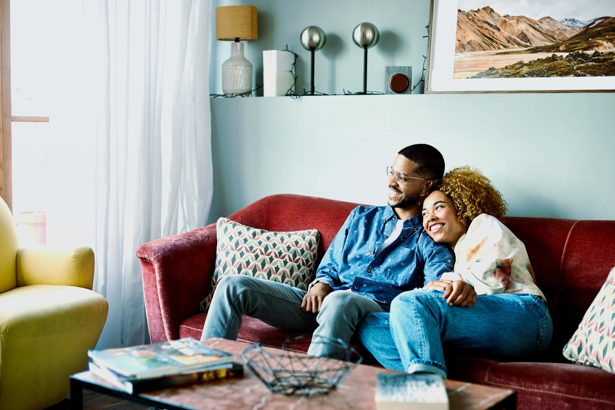 Smiling couple sitting closely together on a red couch in a cozy living room with patterned pillows, a yellow armchair, and a coffee table with books and a decorative wire basket.