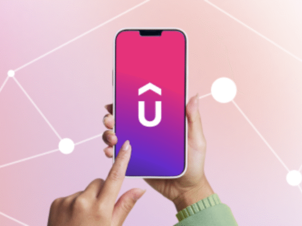 A hand holding a smartphone showing a pink and purple gradient screen with a white uppercase letter U topped by a circumflex accent, while another hand is about to tap the screen, with a pink and white geometric network background.