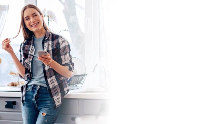 Smiling young woman in a plaid shirt and jeans holding a smartphone and playing with her hair in a bright kitchen with utensils and dried flowers on the counter